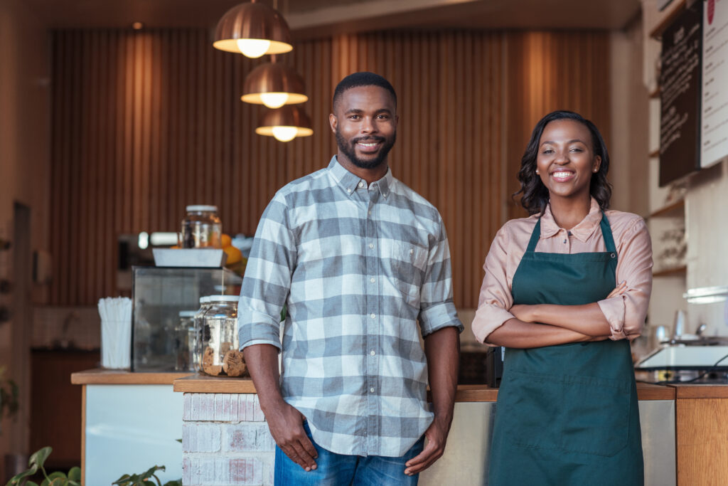 Portrait of two smiling young African entrepreneurs welcomingly togetherin front of the counter of their trendy cafe